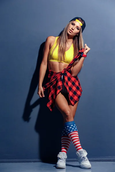 Гламурная сексуальная улыбающаяся красивая молодая женщина модель в спортивной рубашке, шляпе, красных, красных наручных часах, белых кроссовках, леггинсах с флагом США, опираясь на текстурированную стену — стоковое фото