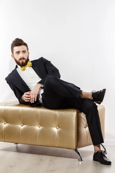 Elegante joven guapo hombre en traje clásico negro. Estudio retrato de moda. Siéntate en el sofá de oro . — Foto de Stock