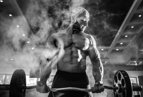 Joven guapo culturista caucásico deportista haciendo ejercicio con una barra en el gimnasio, haciendo el entrenamiento de bíceps, humo —  Fotos de Stock