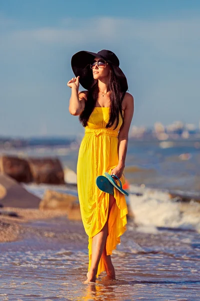 Carefree beautiful fashion brunette woman in beach black hat and long yellow dress flying on the wind walking barefoot by sand of tropical shore.