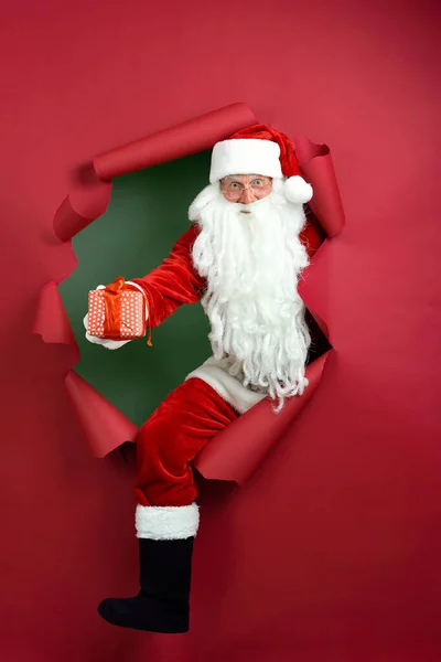 ชายซานต้าถือกล่องของขวัญในมือผ่านรูกระดาษ — ภาพถ่ายสต็อก