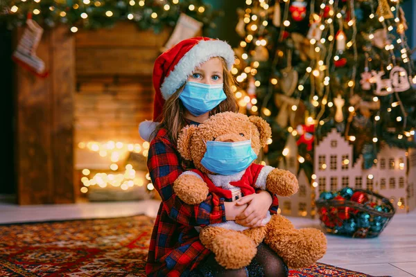 Porträtt av cutel lilla barn flicka i medicinsk mask leka med Taddy björn, Nytt år semester koncept. Konceptet Covid-19 — Stockfoto