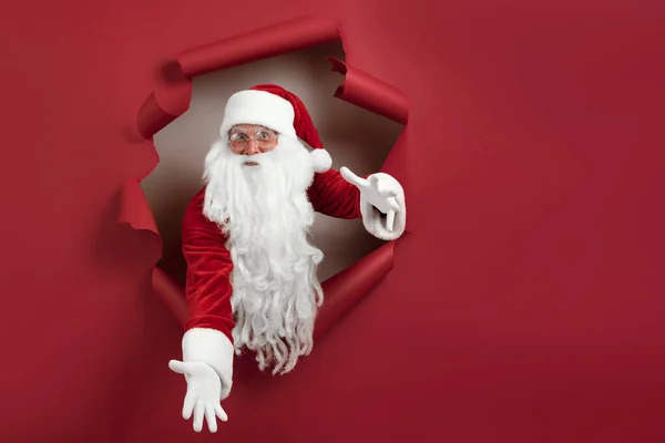 De Kerstman spreidt zijn armen in verschillende richtingen en kijkt emotioneel door een papieren gat. baard man in santa hoed kijken door gat op rood papier — Stockfoto