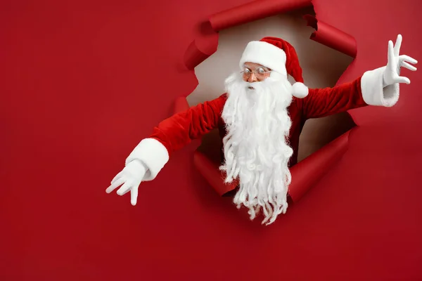 O Papai Noel abriu os braços em direções diferentes e olhou emocionalmente através de um buraco de papel. Homem barbudo em Papai Noel olhando através de buraco em papel vermelho — Fotografia de Stock
