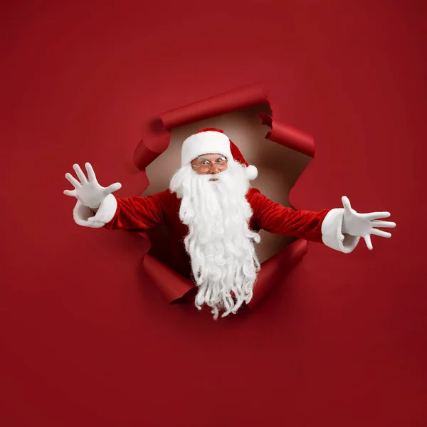 De Kerstman spreidt zijn armen in verschillende richtingen en kijkt emotioneel door een papieren gat. baard man in santa hoed kijken door gat op rood papier — Stockfoto