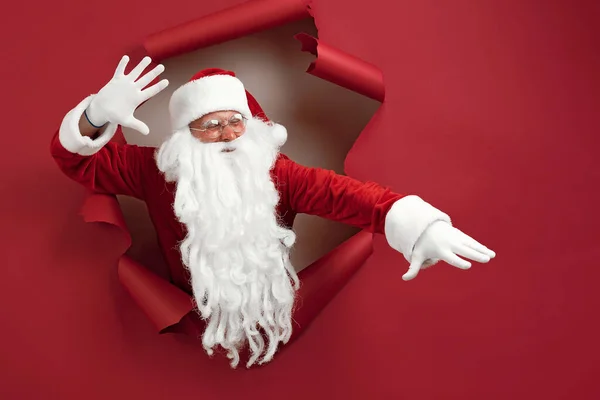 De Kerstman spreidt zijn armen in verschillende richtingen en kijkt emotioneel door een papieren gat. baard man in santa hoed kijken door gat op rood papier — Stockfoto