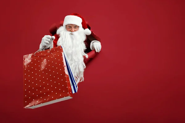 Der Weihnachtsmann hält ein Päckchen in der Hand durch ein Papierloch. Bärtiger Mann mit Weihnachtsmütze blickt durch ein Loch auf rotem Papier — Stockfoto