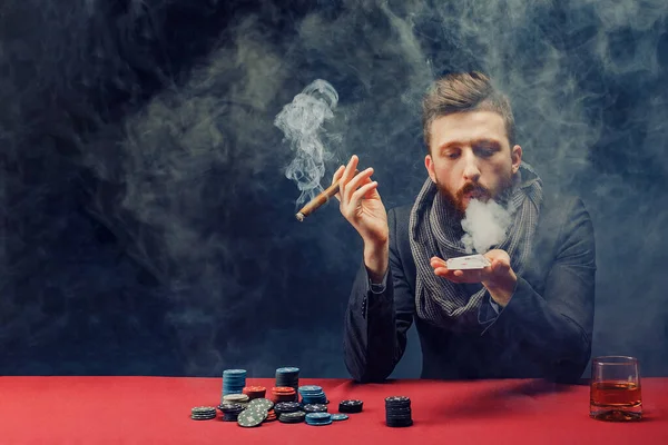 Stylish bearded Man in suit and scarf playing in dark casino, smoking cigar, drink whiskey