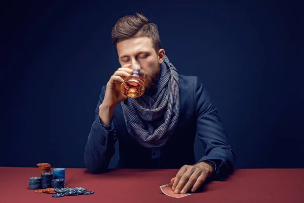 Стильный бородатый мужчина в костюме и шарфе играет в темном казино, курит сигары, пьет виски — стоковое фото