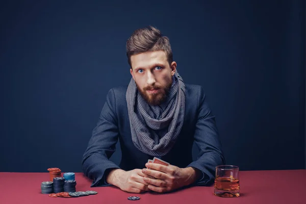 Stylowy brodaty mężczyzna w garniturze i szaliku grający w ciemnym kasynie, palący cygaro, pijący whisky — Zdjęcie stockowe