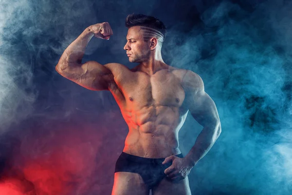 Hombre atlético flexionando los músculos en el estudio sobre fondo oscuro con humo. Culturista fuerte con abdominales perfectos — Foto de Stock