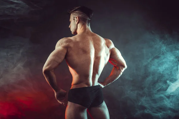 Atlétikai férfi hajlító izmok stúdió sötét háttérrel füst. Erős testépítő tökéletes hasizmokkal — Stock Fotó