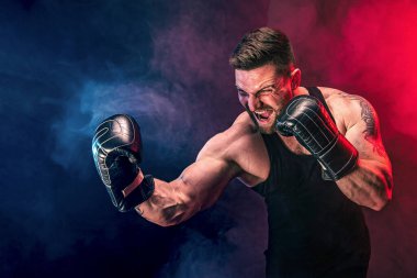 Sakallı sporcu Muay Thai boksörü siyah fanilalı ve boks eldivenli siyah arka planda dumanla dövüşüyor..