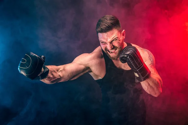 Barbudo tatuado desportista muay thai boxer em camiseta preta e luvas de boxe lutando no fundo escuro com fumaça. — Fotografia de Stock