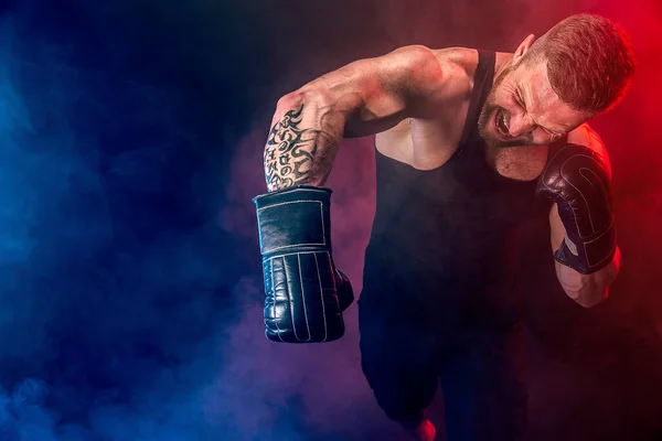 Barbudo deportista tatuado muay thai boxeador en camiseta negra y guantes de boxeo luchando sobre fondo oscuro con humo. — Foto de Stock