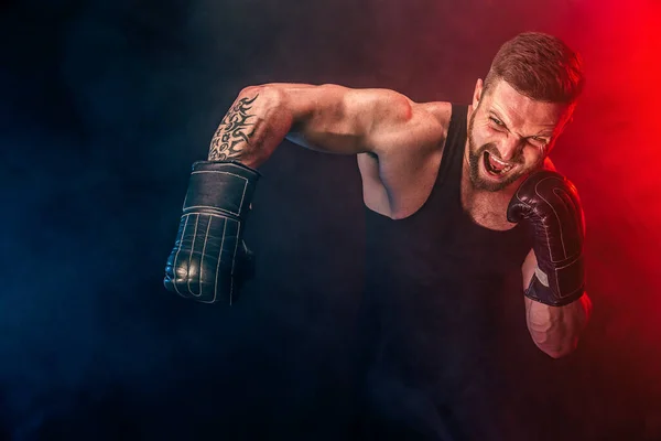 Barbudo deportista tatuado muay thai boxeador en camiseta negra y guantes de boxeo luchando sobre fondo oscuro con humo. — Foto de Stock