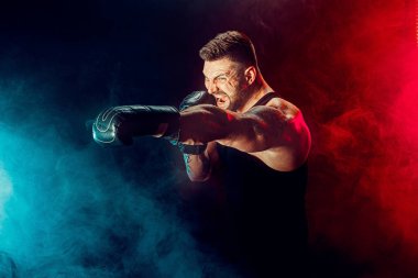 Sakallı sporcu Muay Thai boksörü siyah fanilalı ve boks eldivenli siyah arka planda dumanla dövüşüyor..