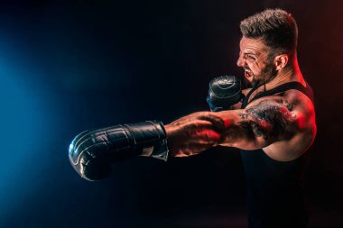 Sakallı sporcu Muay Thai boksörü siyah fanilalı ve boks eldivenli siyah arka planda dumanla dövüşüyor..