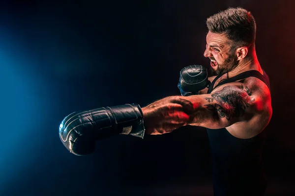 Barbudo deportista tatuado muay thai boxeador en camiseta negra y guantes de boxeo luchando sobre fondo oscuro con humo. — Foto de Stock