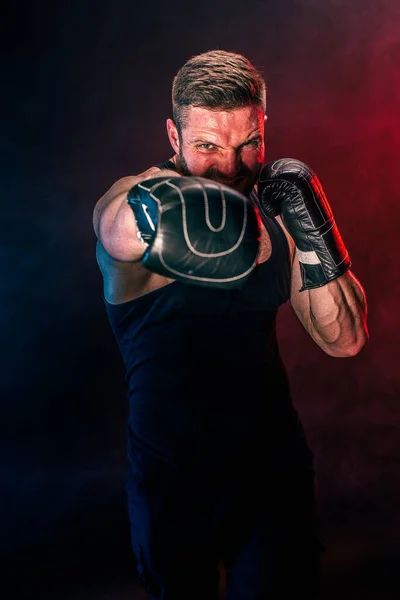 Barbudo deportista tatuado muay thai boxeador en camiseta negra y guantes de boxeo luchando sobre fondo oscuro con humo. — Foto de Stock