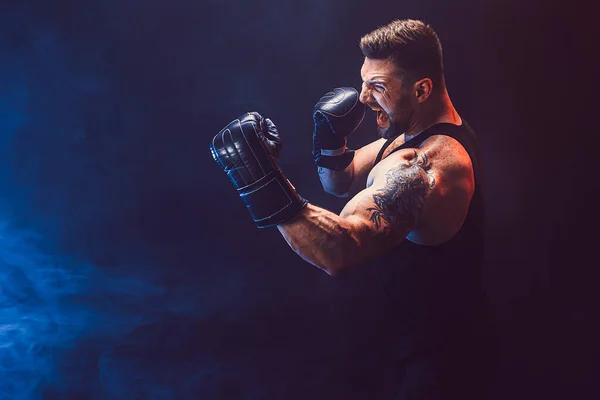 Barbudo deportista tatuado muay thai boxeador en camiseta negra y guantes de boxeo luchando sobre fondo oscuro con humo. — Foto de Stock