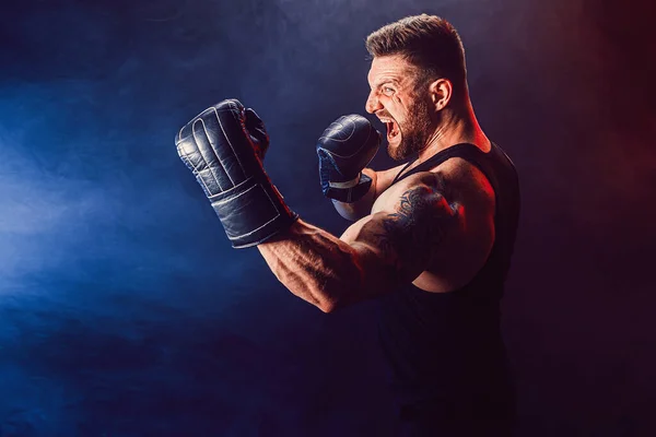 Barbudo deportista tatuado muay thai boxeador en camiseta negra y guantes de boxeo luchando sobre fondo oscuro con humo. — Foto de Stock