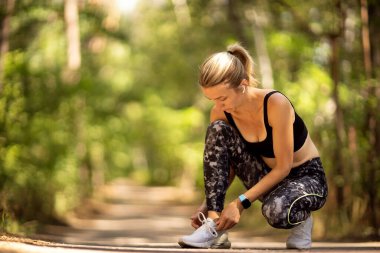 Kadın koşucu spor ayakkabılarını orman yolunda bağlıyor.