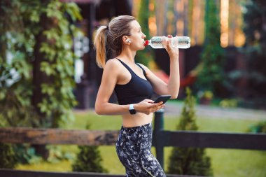 Koştuktan sonra su içen ve akıllı telefon kullanan genç bir kadın.