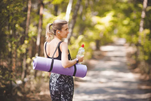 Woman in sport clothes holding a yoga mat and water bottle after