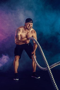 Sakallı sporcu görünüşlü vücut bulucu savaş halatıyla çalışıyor.