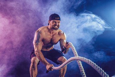 Sakallı sporcu görünüşlü vücut bulucu savaş halatıyla çalışıyor.