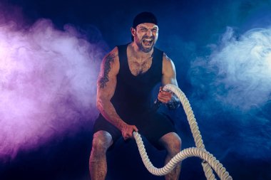 Sakallı sporcu görünüşlü vücut bulucu savaş halatıyla çalışıyor.