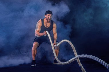 Sakallı sporcu görünüşlü vücut bulucu savaş halatıyla çalışıyor.