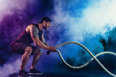 Sakallı sporcu görünüşlü vücut bulucu savaş halatıyla çalışıyor.