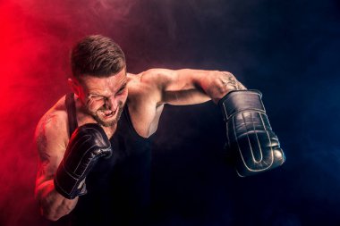 Sakallı sporcu Muay Thai boksörü siyah fanilalı ve boks eldivenli siyah arka planda dumanla dövüşüyor..