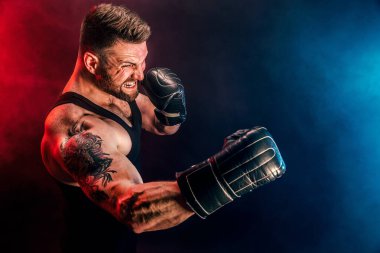 Sakallı sporcu Muay Thai boksörü siyah fanilalı ve boks eldivenli siyah arka planda dumanla dövüşüyor..