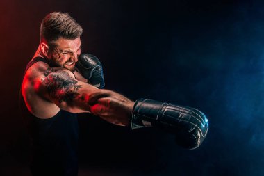 Sakallı sporcu Muay Thai boksörü siyah fanilalı ve boks eldivenli siyah arka planda dumanla dövüşüyor..