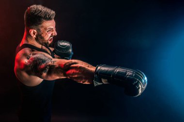 Sakallı sporcu Muay Thai boksörü siyah fanilalı ve boks eldivenli siyah arka planda dumanla dövüşüyor..
