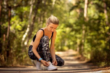 Kadın koşucu spor ayakkabılarını orman yolunda bağlıyor.