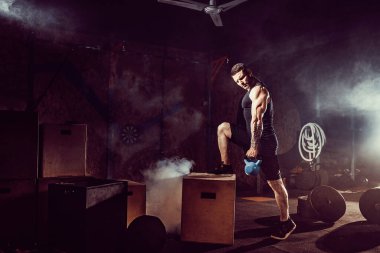 Kaslı, çekici, beyaz sakallı bir adam spor salonunda iki tane kettlebell kaldırıyor. Ağırlık plakaları, halterler ve arka planda lastikler.