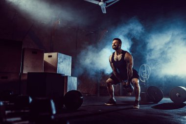 Kaslı, çekici, beyaz sakallı bir adam spor salonunda iki tane kettlebell kaldırıyor. Ağırlık plakaları, halterler ve arka planda lastikler.
