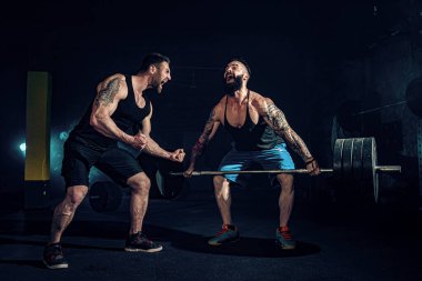 İki kaslı sakallı sporcu spor salonunda antrenman yapıyor.