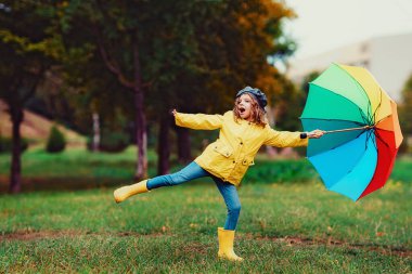 Sonbahar parkında lastik çizmeli, çok renkli şemsiyeli mutlu kız çocuğu.