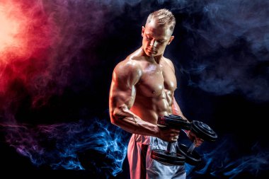 Stüdyoda dambıllarla pazı egzersizi yapan üstsüz bir adam.