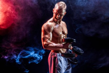 Stüdyoda dambıllarla pazı egzersizi yapan üstsüz bir adam.