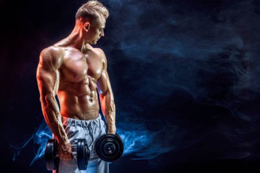 Stüdyoda dambıllarla pazı egzersizi yapan üstsüz bir adam.