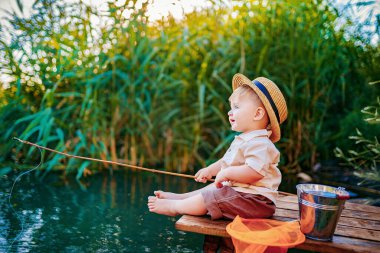 Hasır şapkalı küçük çocuk ahşap bir rıhtımın kenarında oturuyor ve gün batımında gölde balık tutuyor.