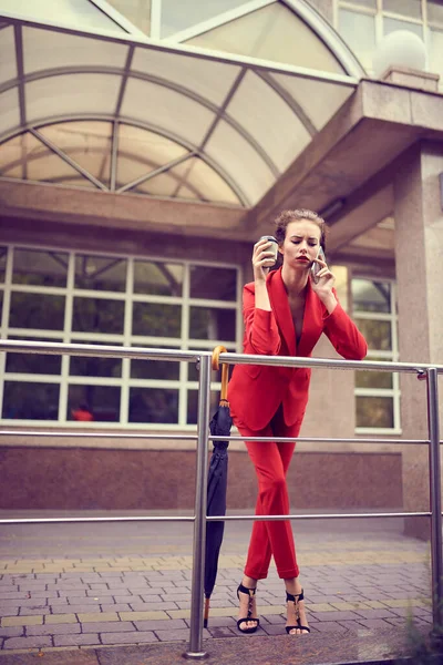 Üzletasszony piros jelmezben mobiltelefon használata irodai közelében, lány böngésző telefon, női menedzser sms okostelefon közelében modern bank épület — Stock Fotó