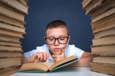 Gözlüklü zeki çocuk iki kitap yığınının arasında oturup kitap okuyor.