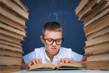 Gözlüklü zeki çocuk iki kitap yığınının arasında oturup kitap okuyor.